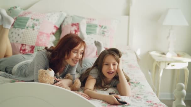 Bela mãe feliz com a filhinha assistindo filme de desenhos animados na TV e rindo enquanto estava deitado na cama em casa pela manhã no quarto acolhedor — Vídeo de Stock