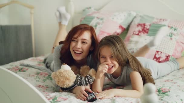 Skrattande glada mor och Söt dotter titta på rolig tecknad film på Tv liggande på sängen hemma på morgonen och ha kul — Stockvideo