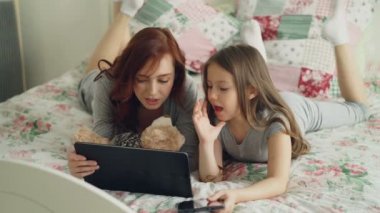 Güzel genç anne ve sevimli kızı gülen ve dijital tablet sabah evde yatakta yatıyordu süre seyir pijama