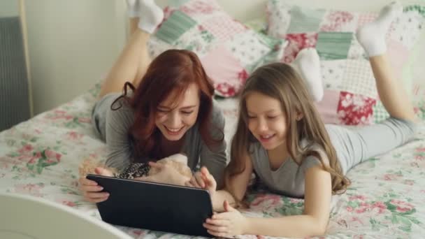 Bella giovane madre e la sua graziosa figlia in pigiama ridendo e guardando in tablet digitale mentre sdraiato sul letto a casa al mattino — Video Stock