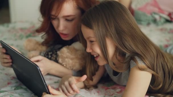 Крупный план привлекательной молодой матери и ее симпатичной дочери в пижаме, смеющейся и смотрящей в цифровые планшеты, лежа на кровати дома утром — стоковое видео
