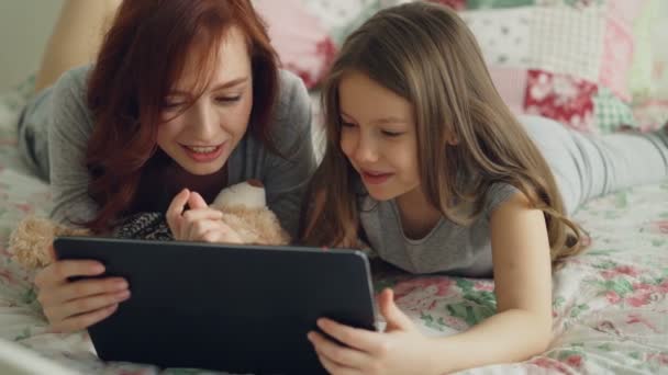 Крупный план улыбающейся милой девушки и молодой матери в пижаме, смеющейся и смотрящей в цифровые планшеты, лежа на кровати дома утром — стоковое видео