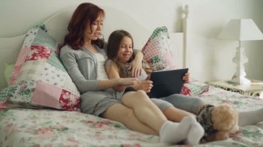 Sevimli kız ve genç annesi gülmek gülümseyen ve sabahları evde yatakta otururken dijital tablet film izlerken