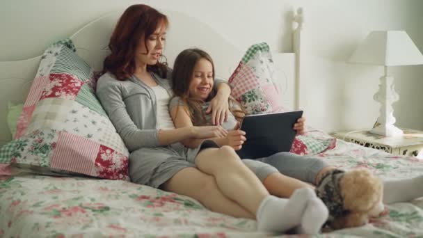 Улыбающаяся милая девушка и ее молодая мама смеются и смотрят кино на цифровой планшет, сидя на кровати дома по утрам — стоковое видео