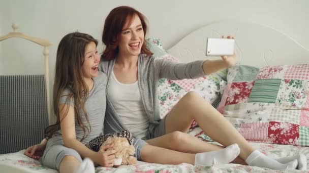 Feliz madre y niña tomando foto selfie con la cámara del teléfono inteligente y divertirse haciendo muecas mientras se sienta en la cama acogedora en casa — Vídeos de Stock