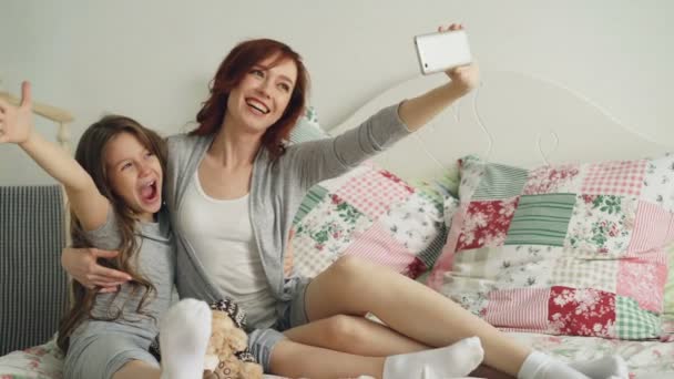 Щаслива мати і маленька дівчинка роблять селфі фотографії з камерою смартфона і отримують задоволення від похмурості, сидячи в затишному ліжку вдома — стокове відео