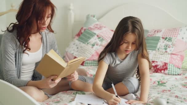 Joven madre ayuda a su pequeña hija linda con la tarea para la escuela primaria. Amar a mamá leyendo un libro y una chica escribiendo notas en copybook mientras están sentados juntos en la cama en casa — Vídeos de Stock