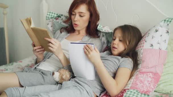 Mãe cuidadosa ajudando sua filhinha fofa com lição de casa para a escola primária. Amante mãe lendo um livro e menina escrevendo notas em livro enquanto sentados juntos na cama em casa — Vídeo de Stock