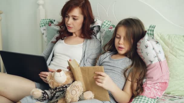 Lilla söta dotter läsa text bokar gör läxor för grundskolan medan hennes kärleksfulla mor surfning och arbetar på digitala tablett sitter tillsammans på mysiga säng hemma — Stockvideo
