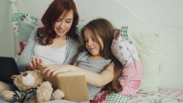 Pequeña linda hija con joven madre cariñosa viendo imágenes divertidas y riendo mientras leen un libro de cuentos de hadas sentados juntos en una cama acogedora en casa — Vídeos de Stock