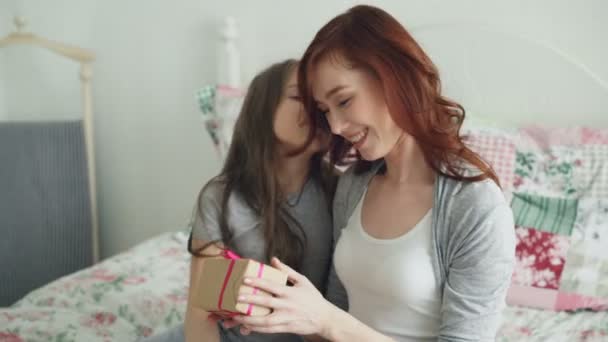 Happy younf mother kissing her cute daughter presenting gift box on celebration sitting on bed in light bedroom at home — Stock Video