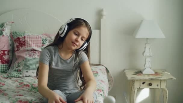 微笑可爱的小女孩在耳机跳舞和移动她的头滑稽, 当坐在床在家在舒适的卧室里 — 图库视频影像