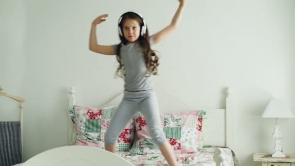 Funny cute little girl in wireless headphones dancing and have fun in holiday morning jumping on bed at home in cozy light bedroom — Stock Video