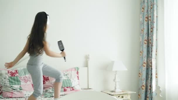 Engraçado bonito menina em fones de ouvido sem fio dançando cantando com pente e se divertir na cama em casa no quarto aconchegante — Vídeo de Stock