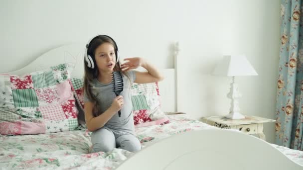 Забавная маленькая девочка в беспроводных наушниках танцует с расчёской и веселится в праздничное утро сидя на кровати дома в уютной спальне — стоковое видео