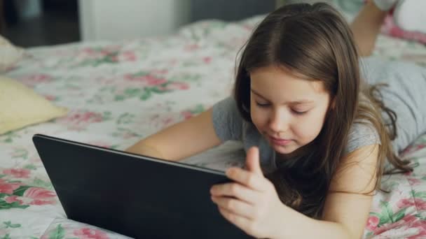 Detailní záběr roztomilé děvčátko pomocí digitálních tabletu a usmívající se přitom ležet v posteli. Dítě mateřská dovolená kreslený film na přenosné zařízení a směje se doma — Stock video