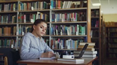 Güzel ciddi beyaz kız öğrenci kitaplığı dizüstü bilgisayar kullanan defterini Özetle aşağı yazma kitaplarla masada oturan