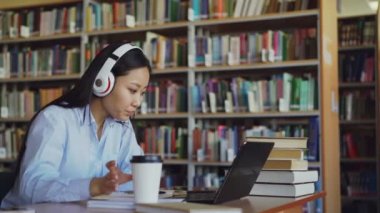 Güzel Asyalı kız öğrencinin kütüphane okul projesi çalışma masasında oturan kulaklıklar müzik dinleme. Dizüstü bilgisayarda yazarak, kahve içme ve defterini içinde yazma genç kız