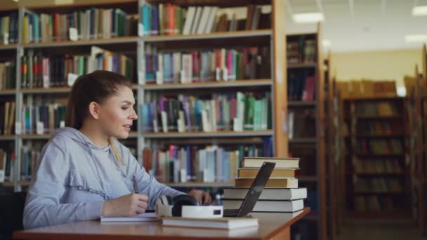 Jeune belle étudiante souriante est assise au bureau dans une grande bibliothèque spacieuse avec une pile de livres. Elle écrit quelque chose dans un copybook, ordinateur portable est en face d'elle — Video