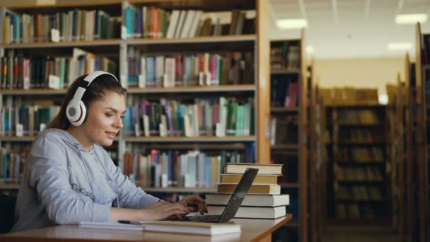 Belle étudiante caucasienne positive avec de gros écouteurs travaillant à table dans une bibliothèque spacieuse devant un ordinateur portable. Elle sourit et envoie des textos — Video