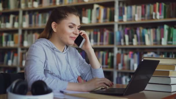 Güzel gülümseyen beyaz kız öğrencinin büyük kütüphane içinde laptop ve onun önünde dijital tablet ile smartphone cep telefonu ile konuşurken masada oturuyor — Stok video