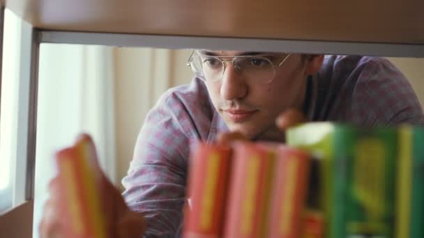 Joven caucásico guapo estudiante masculino está de pie delante de la estantería en la biblioteca universitaria en busca de libro de texto con gafas de poner un libro en otro — Vídeos de Stock