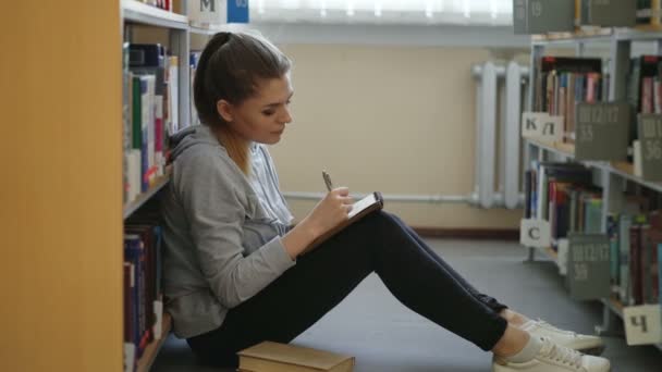 Unga vackra drömmande student tjej sitter på golvet i svagt bibliotek bland bokhyllor skriva ner sammansättning i häfte tittar upp på taket — Stockvideo