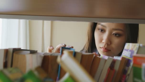 Красивая азиатская вдумчивая студентка ищет необходимую книгу на книжной полке в большой библиотеке, открывая ее и улыбаясь довольной — стоковое видео