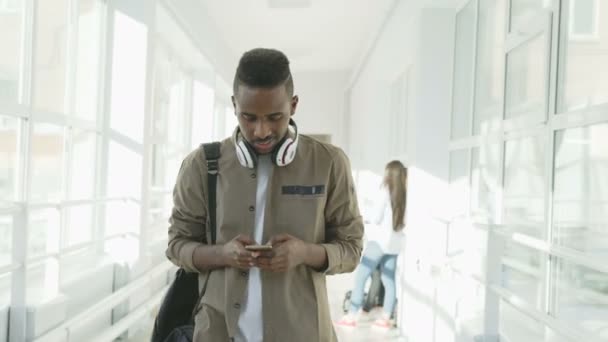 Симпатичный афро-американец, студент с большими белыми наушниками, прогуливающийся по длинному светлому коридору колледжа, держит смартфон и пишет кому-то — стоковое видео