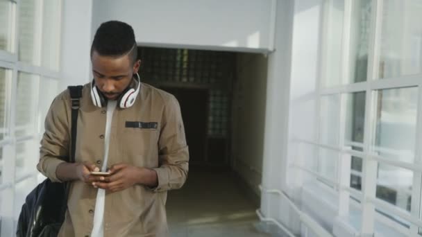 Stilig afro-american manlig student med stora vita hörlurar gå i länge svagt korridor av college hålla smartphone SMS någon — Stockvideo