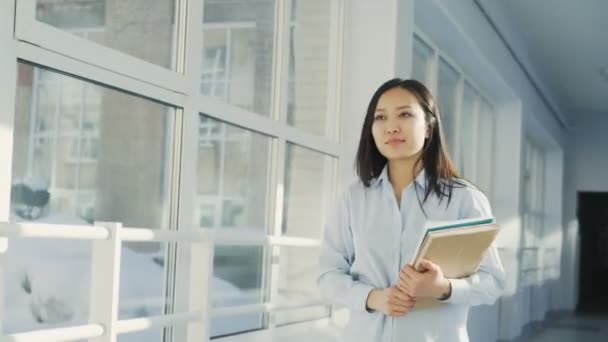 Giovane bella ragazza asiatica che cammina lungo il corridoio nel college con libri di testo e documenti notando gli amici, salutandoli e sorridendo, ragazzo afro la sta guardando — Video Stock