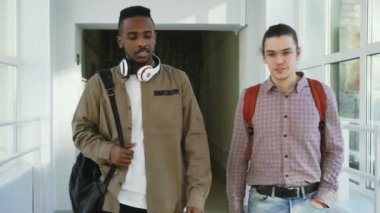 İki genç yakışıklı çok etnik gruptan oluşan arkadaş beyaz cam gibi koridor ayakta kızlar tarafından geçen ve onlara el sallayarak konuşuyor üniversitede aşağı yürüyüş