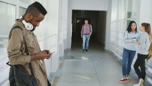 Gruppo di studenti è in piedi in corridoio ragazza stanno parlando ragazzo afro è in possesso di smartphone bello studente viene agitando mano alle ragazze e fare saluto al suo amico — Video Stock