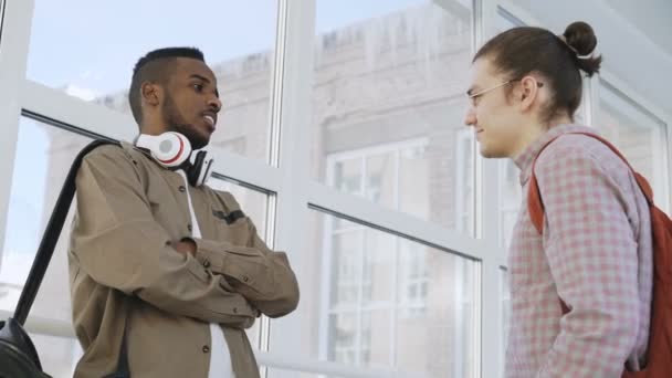 Dois jovens bonitos estão em pé no grande corredor da faculdade vítreo comunicando de forma positiva e sorrindo. Estudante afro está expalando algo para caucasiano — Vídeo de Stock