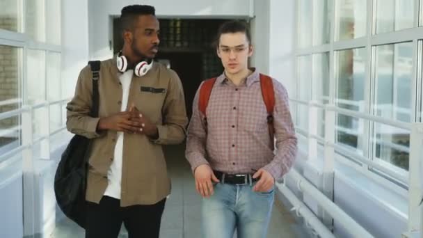 Kaukasische hipster en Afro-Amerikaanse studenten gaan op college grote glazig gang bespreken iets handen zwaaien naar vrouwelijke klasgenoten en zet op — Stockvideo