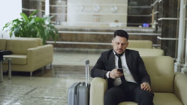 Plan panoramique d'un homme d'affaires concentré utilisant un smartphone assis sur un fauteuil à l'intérieur d'un hôtel de luxe avec des bagages près de lui — Video