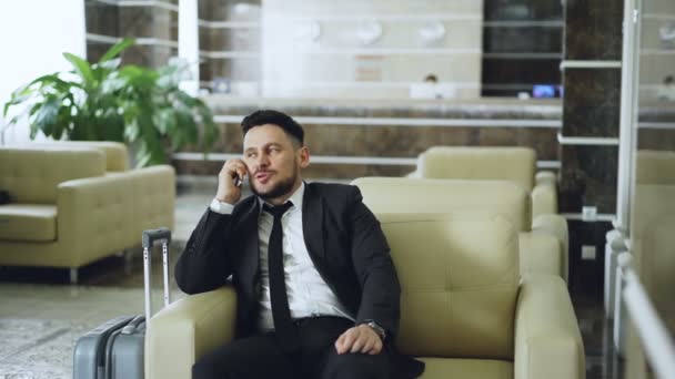 Pan tiro de empresário confiante falando telefone celular, enquanto se senta em poltrona em hotel de luxo com bagagem perto dele. Conceito de viagem, negócios e pessoas — Vídeo de Stock
