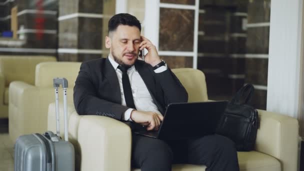 Tilt up of confident businessman talking mobile phone and using laptop computer while sits on armchair in luxury hotel with luggage near him. Travel, business and people concept — Stock Video