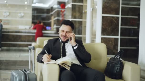 Panorámica de alegre empresario confiado hablando de teléfono móvil sonriendo y escribiendo notas en bloc de notas mientras se sienta en el sillón en la recepción del hotel. Concepto de viajes, negocios y personas — Vídeos de Stock