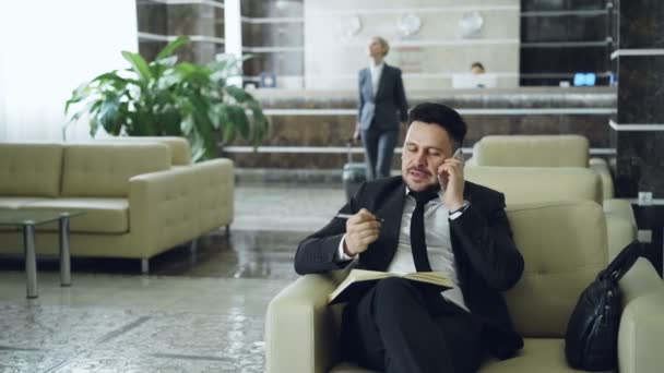 Пан снимок красивого бородатого бизнесмена, сидящего в кресле и разговаривающего по мобильному телефону с блокнотом, в то время как деловая женщина с багажом, идущая через вестибюль отеля со стойки регистрации — стоковое видео