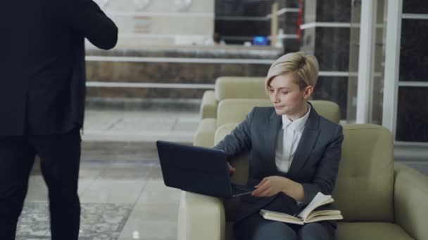 Блондинка-бизнесвумен сидит в кресле с блокнотом и ноутбуком, в то время как бизнесмен с багажом идет через вестибюль отеля на стойку регистрации — стоковое видео