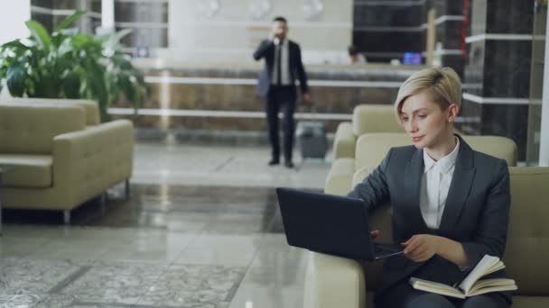 Blond kobieta siedzi w fotelu z Notatnika i laptop komputer podczas biznesmen z bagażem spacer hotelowym lobby z recepcją — Wideo stockowe