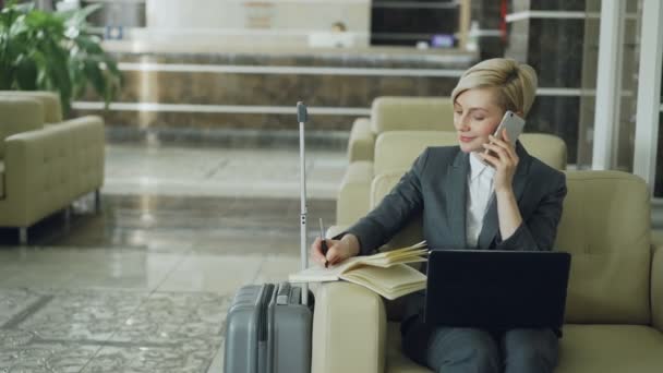 Panorera bilden av blond upptagen affärskvinna sitter i fåtölj i hotellobbyn prata mobiltelefon, skriver i anteckningar och använder bärbar dator. Affärer, resor och människor-konceptet — Stockvideo