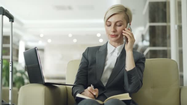 Blonde busy businesswoman sitting in armchair in hotel lobby talking mobile phone, writing in notepad and using laptop computer. Business, travel and people concept — Stock Video
