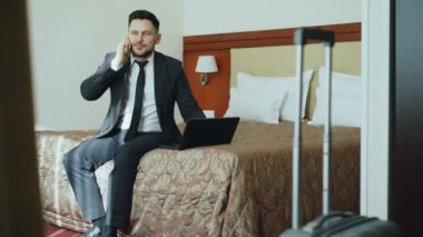 Dizüstü bilgisayarda çalışan ve yatakta otel odasında oturma sırasında cep telefonu konuşurken elbiseli gülümseyen iş adamı yukarı eğimli. İş, seyahat ve insanlar kavramı