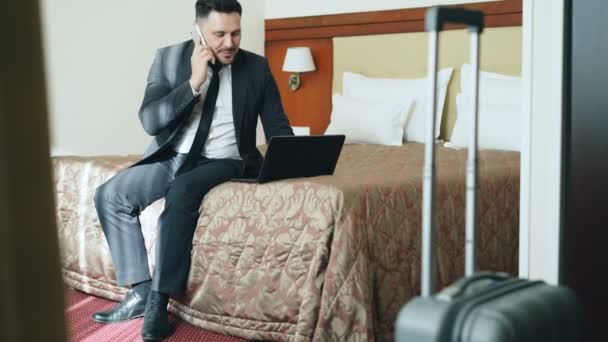 Наклон занятого бизнесмена в костюме, работающего на ноутбуке и разговаривающего по мобильному телефону, сидя на кровати в гостиничном номере. Концепция бизнеса, путешествий и людей — стоковое видео