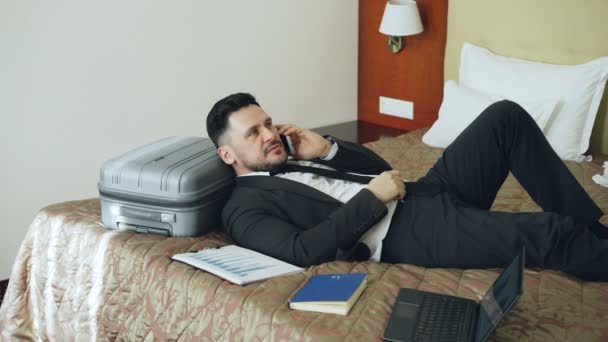 PAN of confident businessman talking mobile phone while lying on bed in hotel room. Путешествия, бизнес и люди — стоковое видео