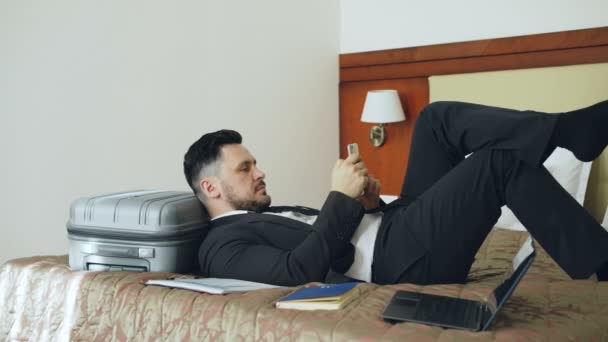 Jovem empresário deitado na cama e na mala usando smartphone enquanto descansa após a chegada ao quarto de hotel. Conceito de viagem, negócios e pessoas — Vídeo de Stock