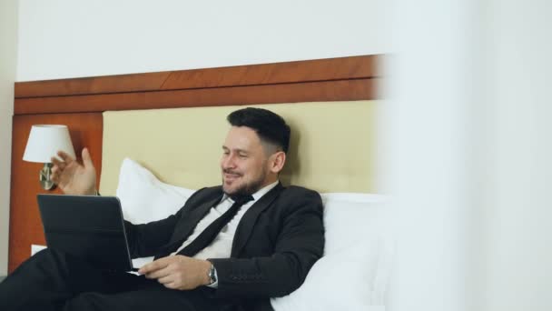 Счастливый бизнесмен разговаривает с семьей через онлайн видео чат, используя ноутбук, лежащий на кровати в гостиничном номере. Путешествия, бизнес и люди — стоковое видео