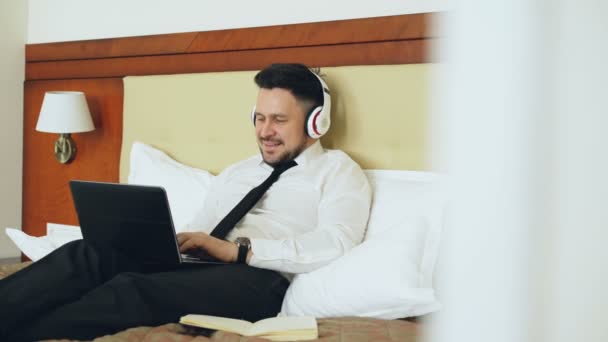 Happy businessman in headphones working at laptop computer and listening music smiling while lying in bed at hotel room. Travel, business and people concept — Stock Video